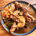 A plated dish of cooked chicken pieces in a brown sauce with a colorful vegetable topping, served on a wooden table next to a white bowl of white rice.