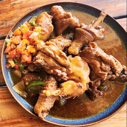 A plated dish of cooked chicken pieces in a brown sauce with a colorful vegetable topping, served on a wooden table next to a white bowl of white rice.