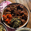 A close-up of a plated meal on a wooden table, featuring a bowl of stewed meat in a dark brown sauce with side vegetables, a bowl of white rice, and another partial dish at the lower left.