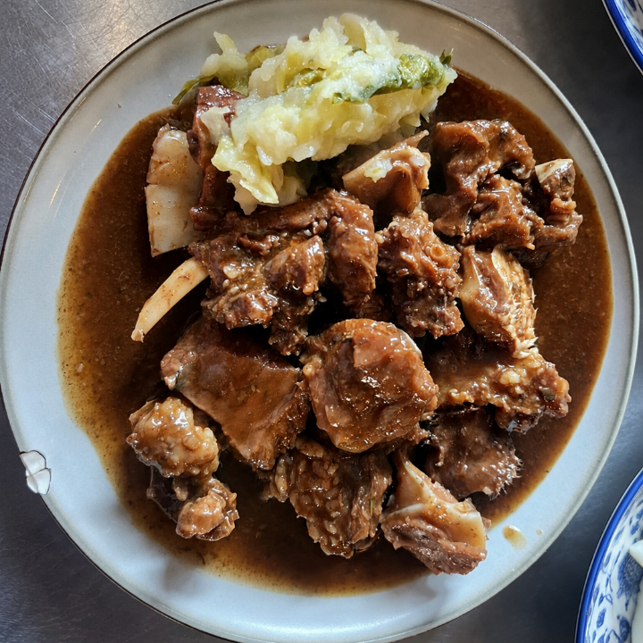 A top-down view of a plate of braised meat with brown sauce and a side of pale cabbage on a stainless steel table.