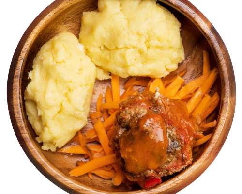 A close-up overhead shot of a wooden bowl containing mashed yellow potatoes, shredded orange carrots, and a piece of meat topped with orange-red sauce, isolated on a white background.