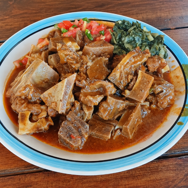 A plate of traditional bone stew with chunks of meat and bone in a rich reddish-brown sauce, served with chopped tomatoes and cooked greens on a blue-rimmed white plate, set on a wooden table.