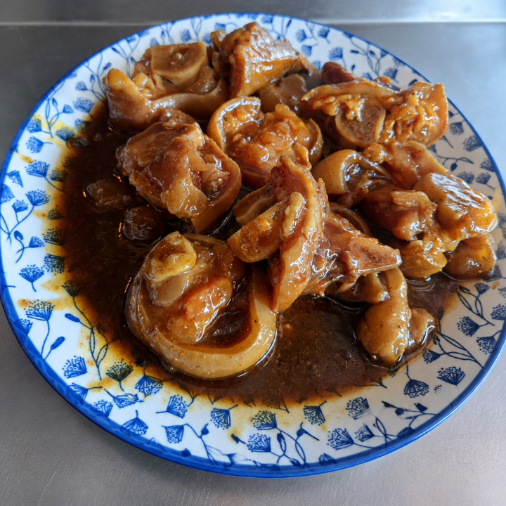 A plate of braised or stewed meat pieces in a dark brown sauce, served on a blue and white patterned ceramic plate against a stainless steel background.