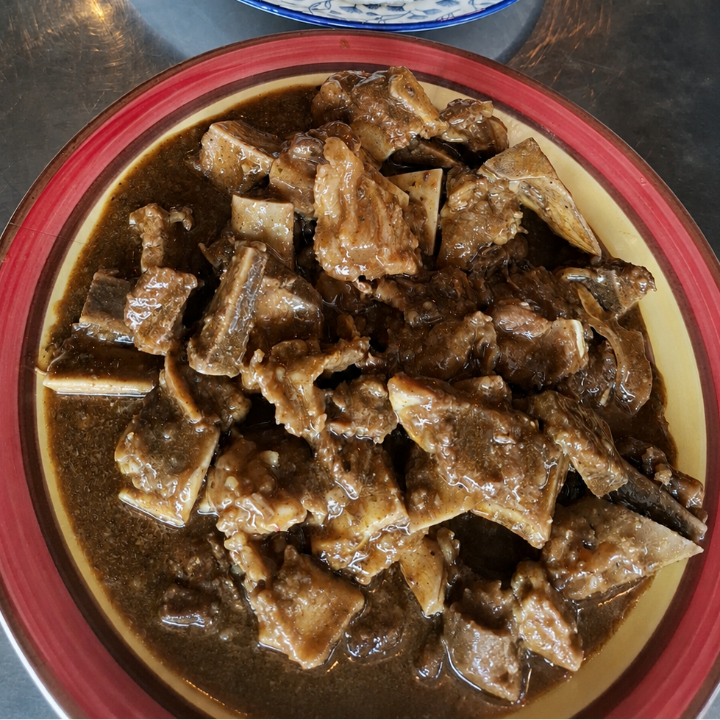 A close-up of a meal on a dark metallic table, featuring a plate of white steamed rice or dough and a bowl of braised meat chunks in a brown sauce.