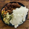 A top-down view of a black plate containing mashed potatoes, braised meat, coleslaw, and a side of mixed vegetables, set on a rustic wooden table.